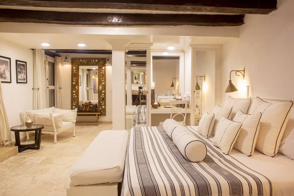 A well-lit bedroom with a large bed, a desk, and a chair. There are curtains on the windows and a mirror on the wall.