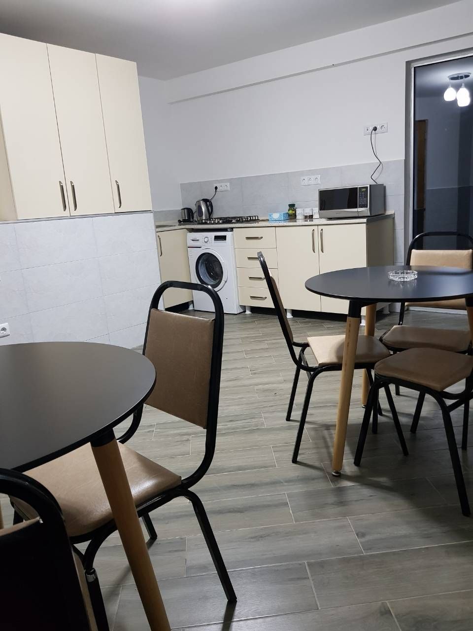 Een praktische keuken met een magnetron, waterkoker en een wasmachine. Er staan twee eettafels met stoelen.