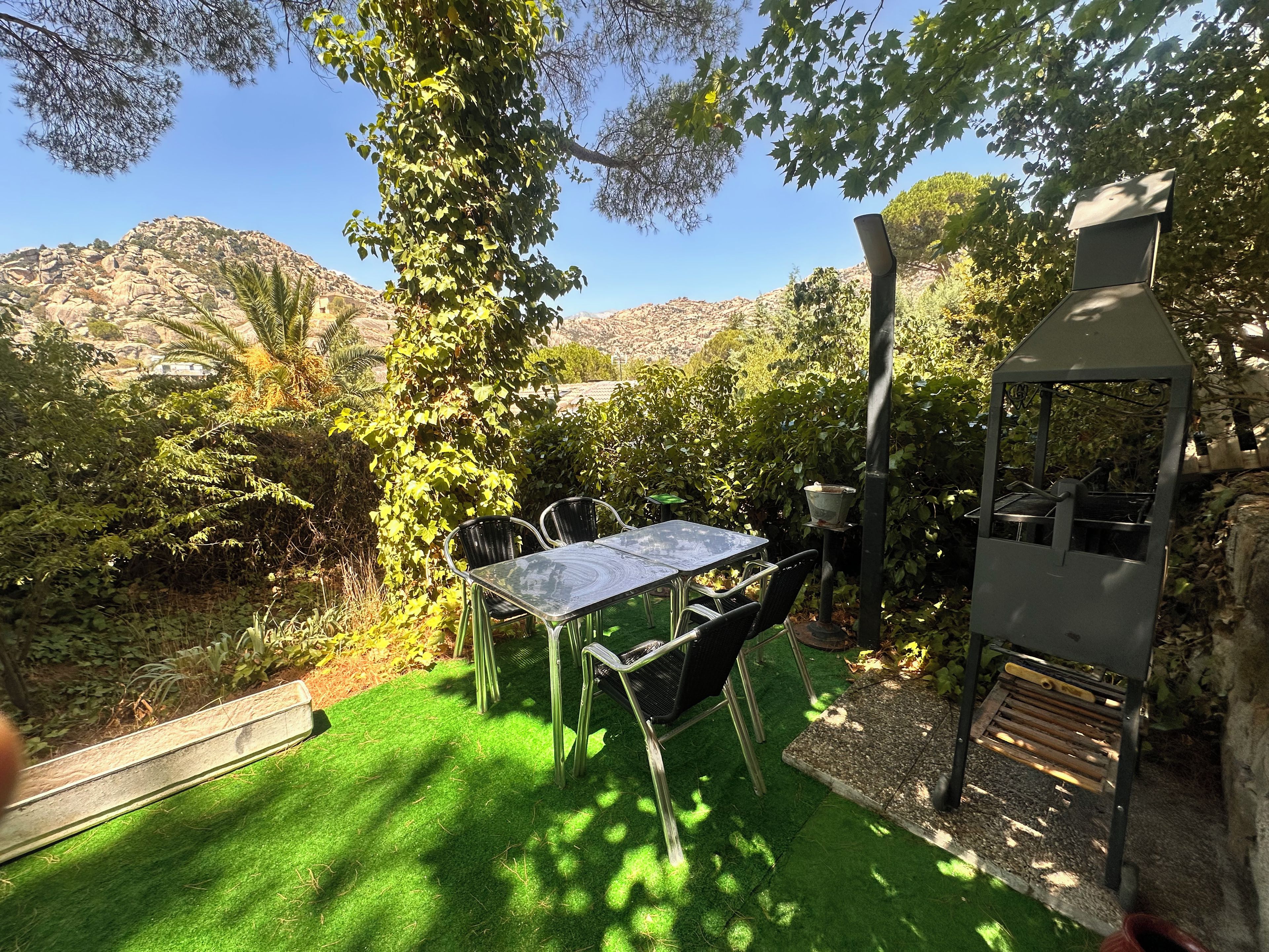 A patio with a table and chairs, surrounded by greenery and a view of hills.