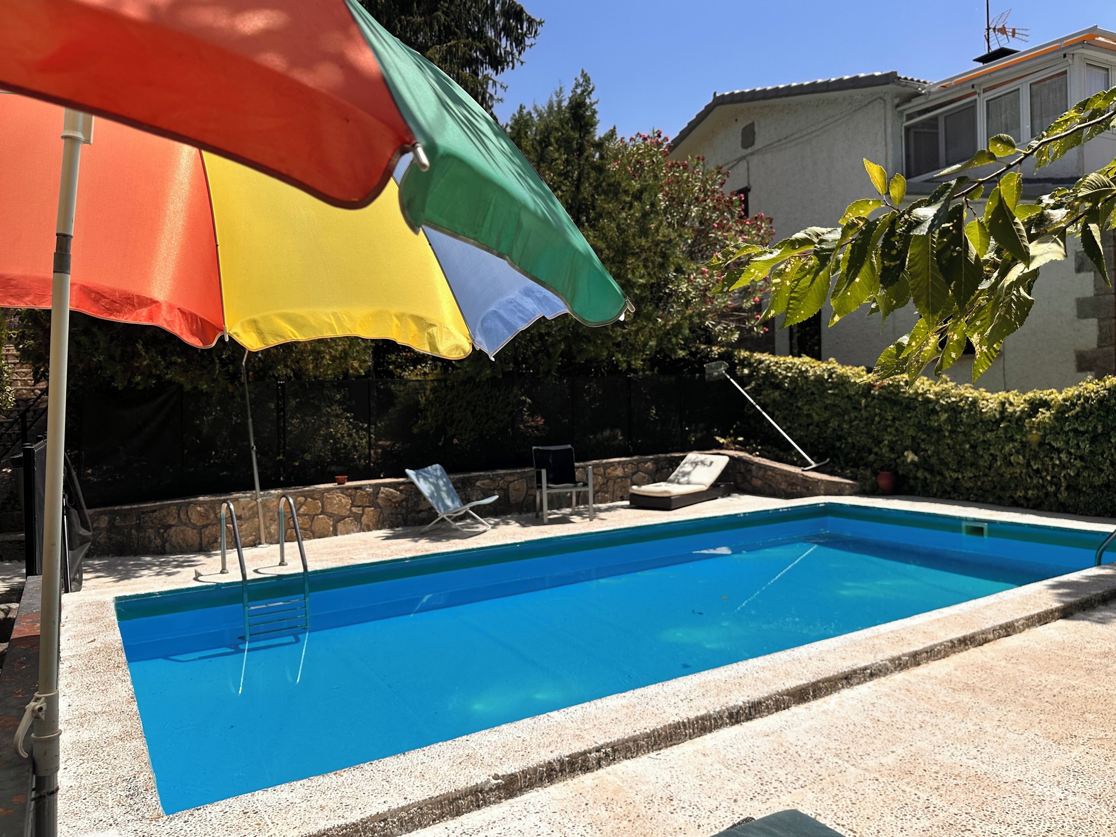A swimming pool with a clear blue water surface, surrounded by a stone wall and a hedge. There are lounge chairs and a table under a colorful umbrella.