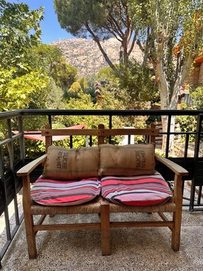 Un banco de madera con cojines a rayas y un cojín de yute, situado en un balcón con vistas a un paisaje montañoso.