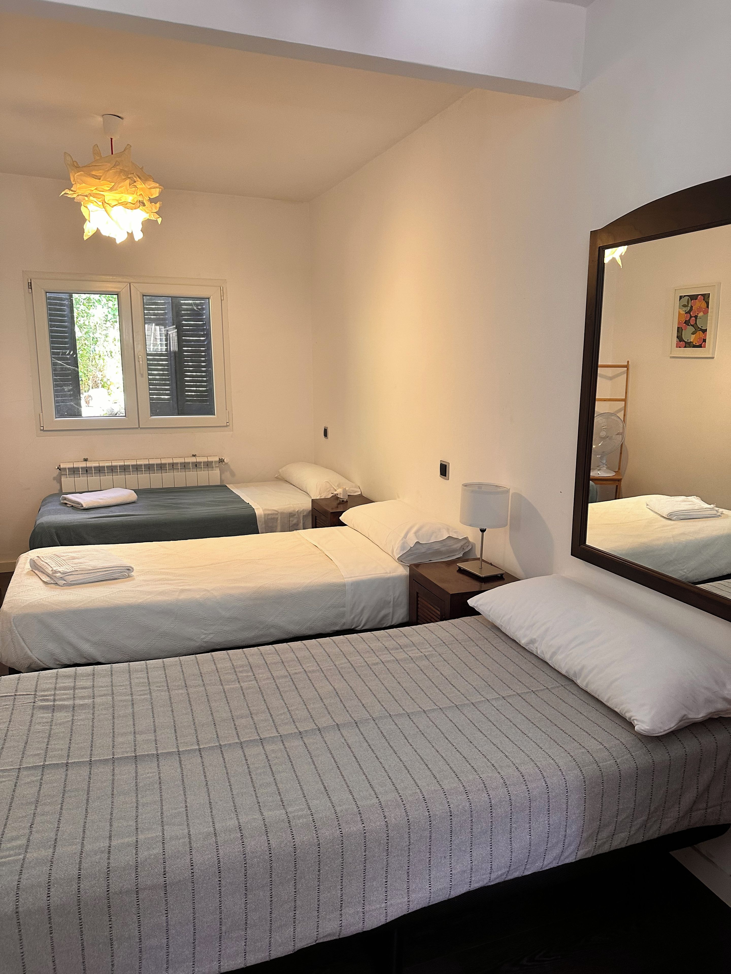 A hotel room with two beds, a radiator, a window with shutters, a bedside table with a lamp, and a mirror.