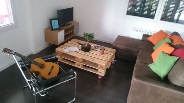 Un salon avec une table basse en bois, une guitare, un téléviseur posé sur un meuble, un canapé agrémenté de coussins colorés et une plante en pot.