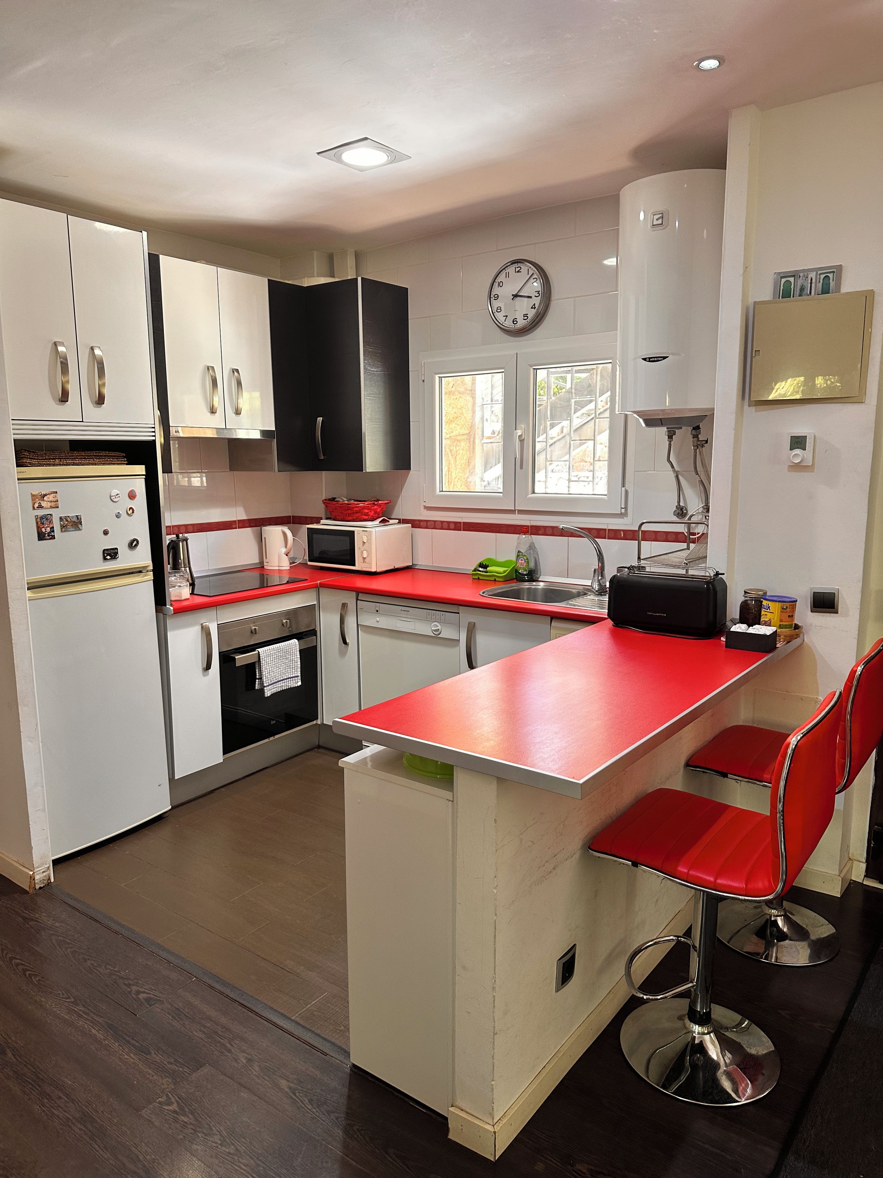 Una cucina con mobili bianchi, un piano di lavoro rosso, una sveglia e uno scaldabagno.