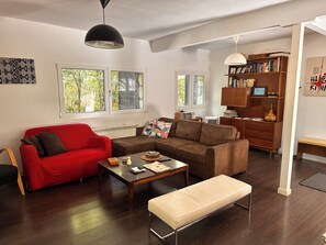 A living room with a red sofa, a brown sofa, a wooden coffee table, and a bench. There are two windows with a view of trees, a bookshelf, and a radiator.