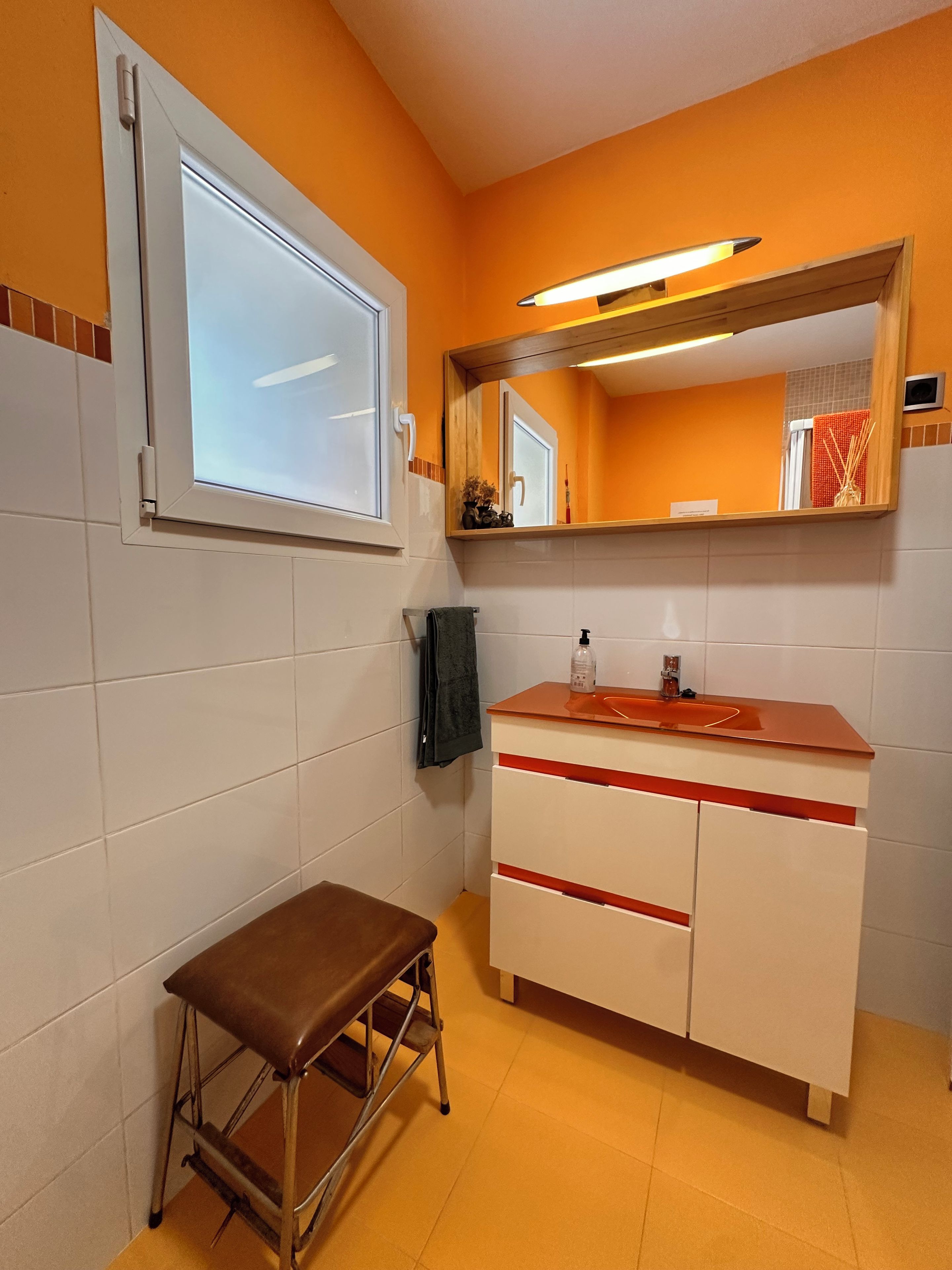 A bathroom with a white vanity, a mirror, a window, and a bench.