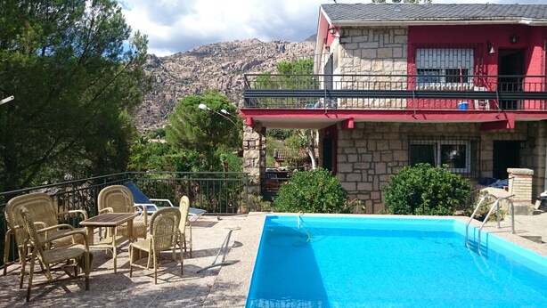 Zona de piscina con sillas de mimbre, un edificio de piedra con fachada roja y montañas al fondo.