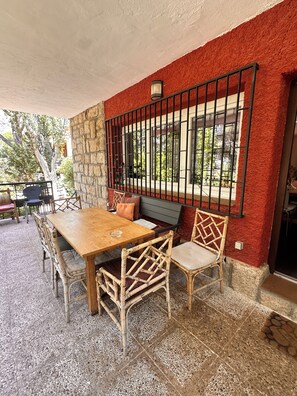 Un espacio exterior para comer con una mesa de madera, sillas de mimbre, una pared de piedra y una ventana con rejas.