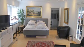 A bedroom with a bed, a TV, a plant, a painting, a chair, and a door. - Guesthouse Sharon Suite (Aarhus)