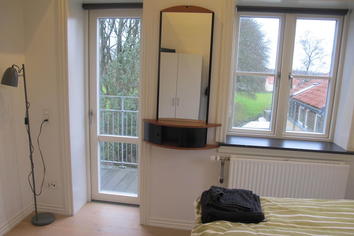 Un dormitorio con cama, ventana con vista a un jardín, radiador y puerta que da a un balcón.