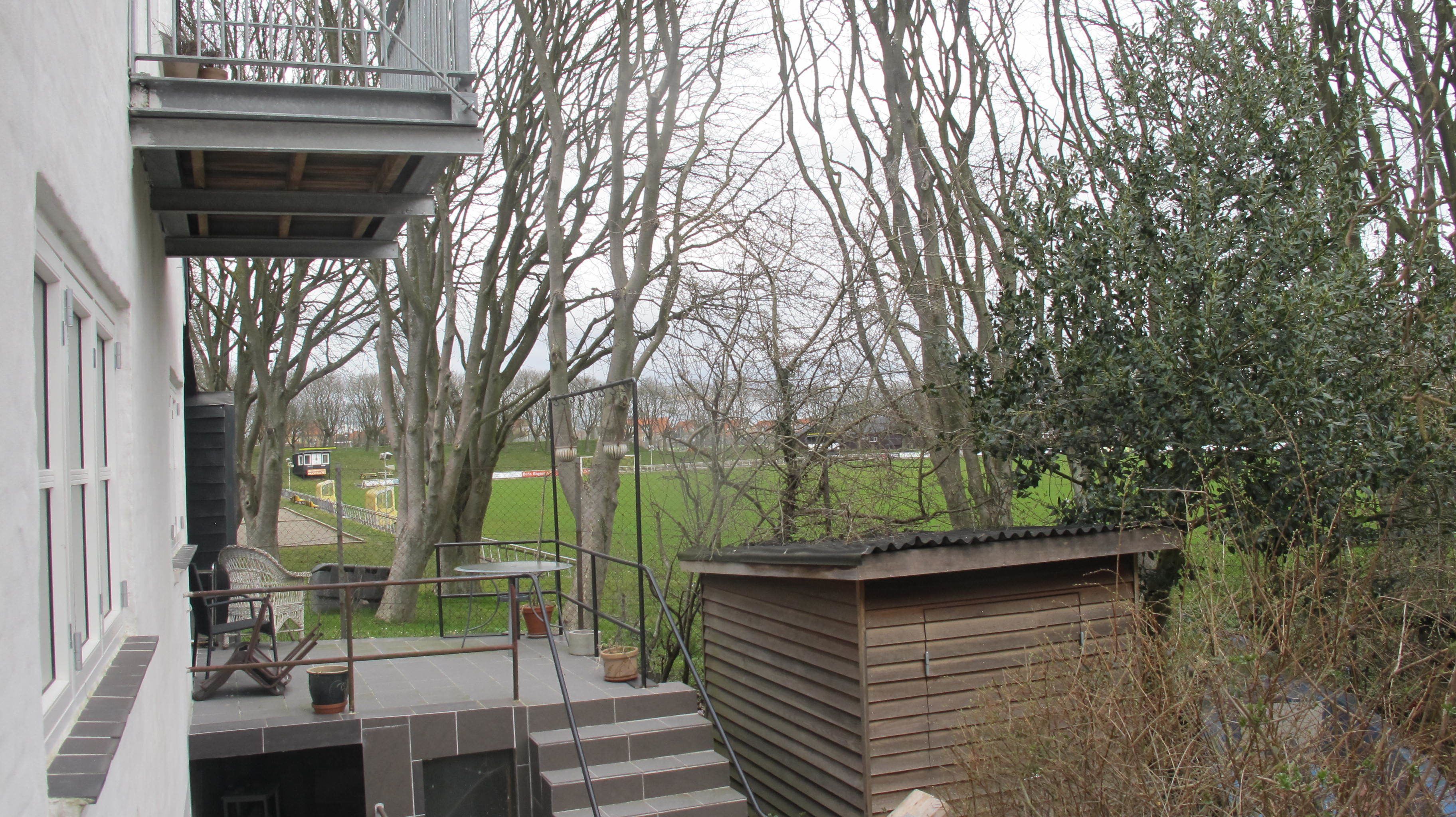 Un quartier résidentiel avec un escalier menant à un balcon, une petite construction en bois et une vue sur un parc arboré et un bâtiment situé au loin.