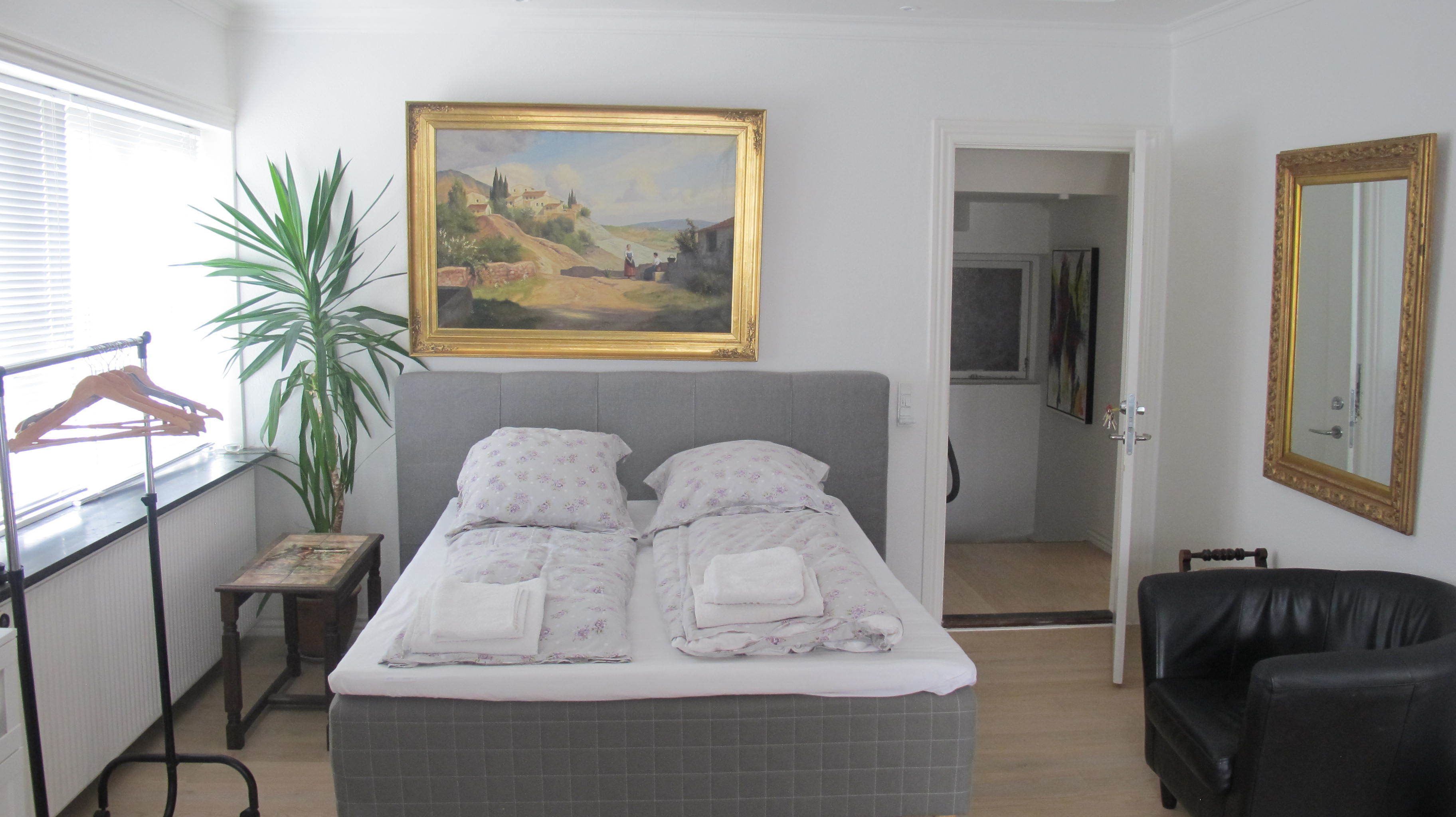 A bedroom with a bed, a painting, a mirror, a chair, and a plant.