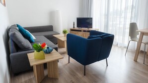 A modern living room with a grey sofa, a blue armchair, a wooden coffee table with a vase of flowers, a flat-screen TV on a wooden stand, and a dining area with a table and chairs. - Pebble Beach Holiday Apartments (Limassol)