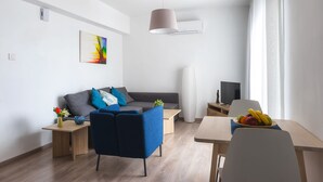 A modern living room with a sofa, a coffee table, a dining table with fruit, a TV, and a large window with curtains. - Pebble Beach Holiday Apartments (Limassol)