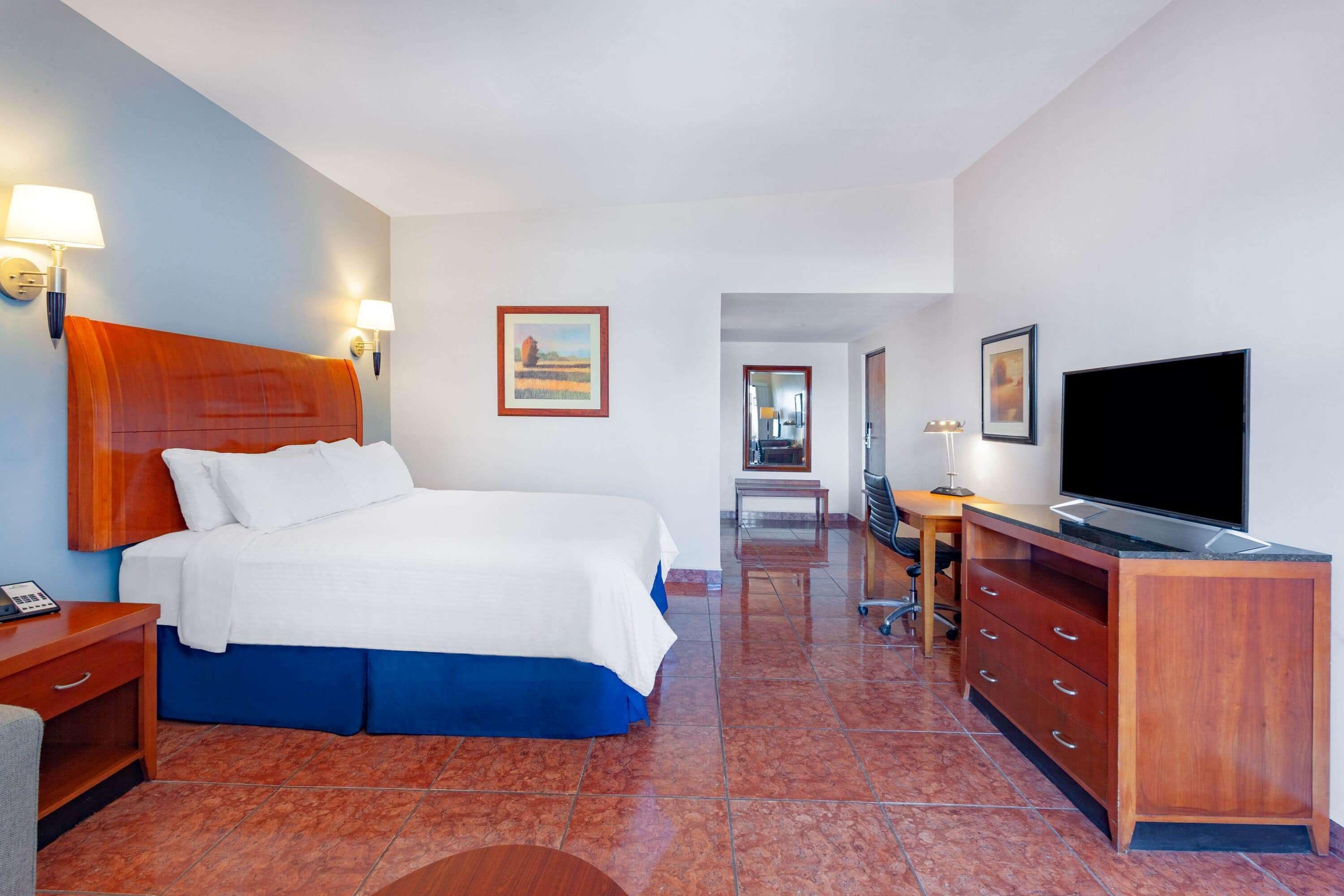 A hotel room with a large bed, a desk with a flat-screen TV, a dresser, and a mirror.