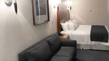 Habitación de hotel con un sofá gris, una cama con ropa de cama blanca, dos lámparas de pared y un cuadro en la pared.