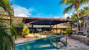 A swimming pool with a stone deck, a covered seating area, and a thatched-roof structure.