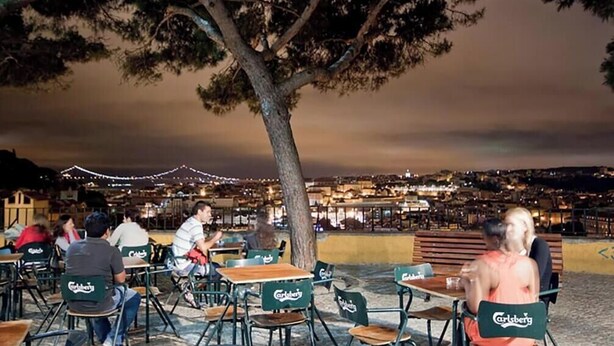 An outdoor café with tables and chairs, a tree in the center, and a cityscape in the background.