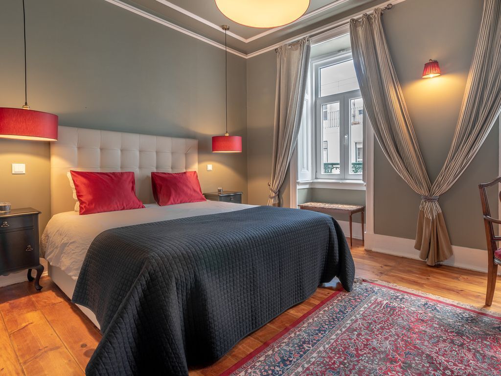 Een slaapkamer met een groot bed, rode kussens, een donge sprei, een rode lamp, een raam met gordijnen en een kamerbreed tapijt met een patroon.