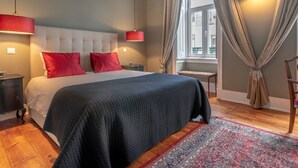 A bedroom with a large bed, red pillows, a dark quilt, a red lamp, a window with curtains, and a patterned rug.