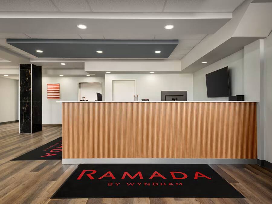 Een receptiegedeelte met een houten receptiebalie, een flatscreen-tv en een zwarte deurmat met 'RAMADA BY WYNDHAM' erop.