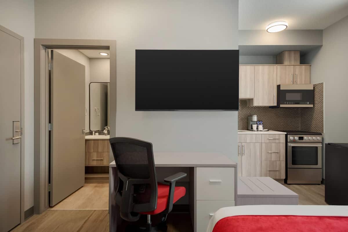 Habitación de hotel moderna con cama, escritorio y silla, televisor de pantalla plana, kitchenette con microondas y horno, y baño con espejo y lavamanos.