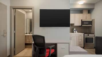 Habitación de hotel moderna con cama, escritorio y silla, televisor de pantalla plana, kitchenette con microondas y horno, y baño con espejo y lavamanos.