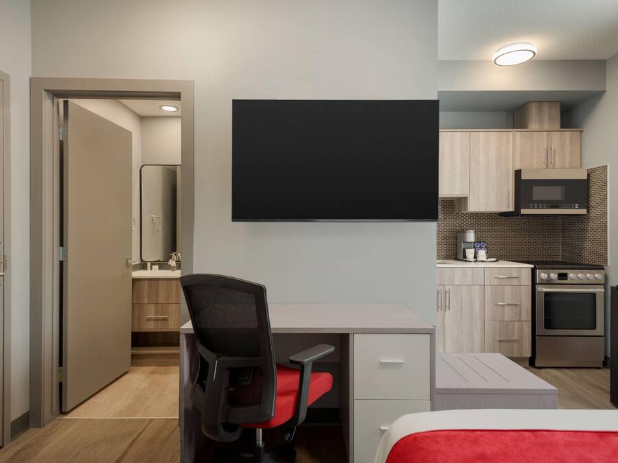 Habitación de hotel moderna con cama, escritorio y silla, televisor de pantalla plana, kitchenette con microondas y horno, y baño con espejo y lavamanos.