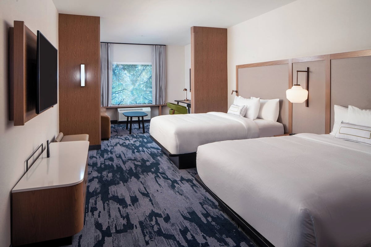 A hotel room with two beds, a desk, a chair, a TV, and a window with a view of trees.