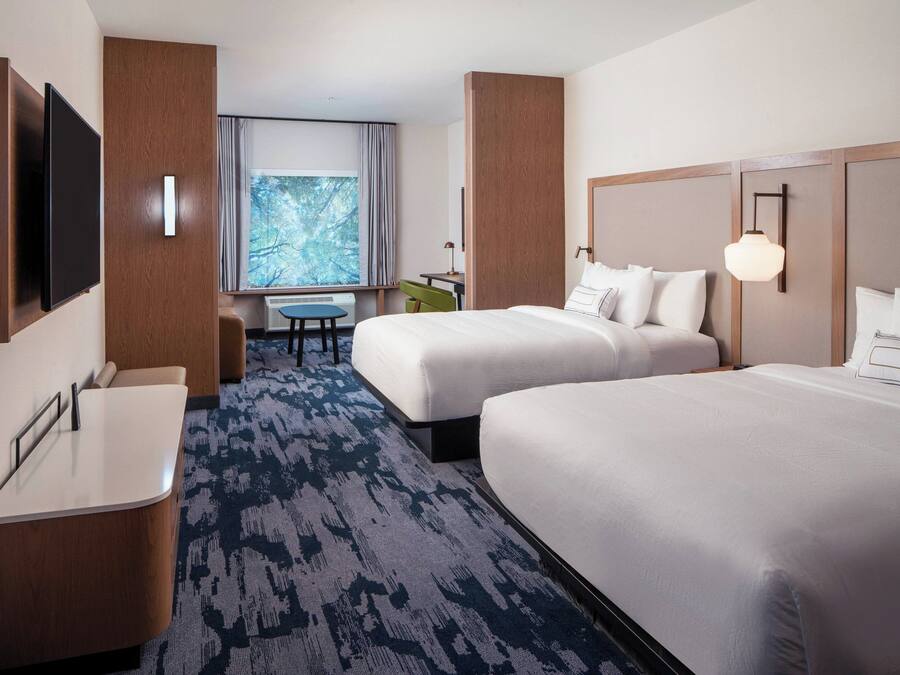 Ein Hotelzimmer mit zwei Betten, einem Schreibtisch, einem Sessel, einem Fernseher und einem Fenster mit Blick auf Bäume.