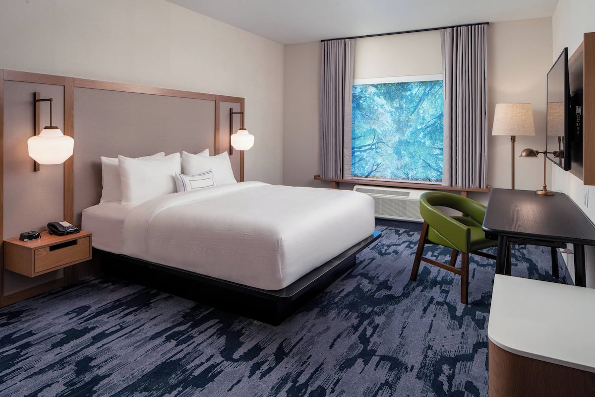 A hotel room with a large bed, bedside lamps, a desk with a chair, a window with curtains, and a flat-screen TV.