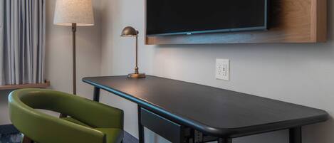 A modern hotel room with a flat-screen TV mounted on the wall, a desk with a lamp, and a green chair.