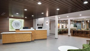 A modern reception area with a wooden reception desk, a large abstract wall art piece, and a seating area with green upholstered chairs. - Fairfield by Marriott Inn & Suites Pensacola Pine Forest (Pensacola)