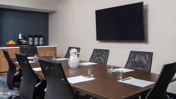 A conference room with a large table, six chairs, a television mounted on the wall, and a small kitchenette area with a fruit bowl and beverage dispensers.