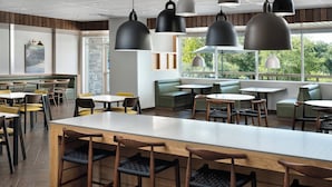 Interior de un restaurante con mesas y sillas de madera, una barra central y amplios ventanales con vistas a zonas verdes.