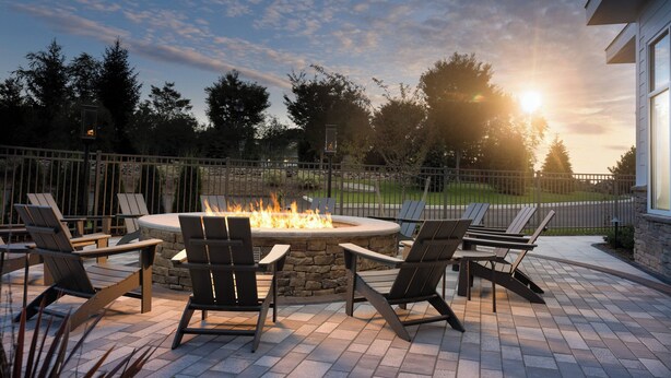 Un área de patio con un hogar de piedra rodeado de sillas Adirondack, un muro de piedra y vistas a un vecindario residencial al atardecer.