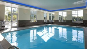 Una piscina interior con paredes de vidrio transparente, suelo de baldosas y escalera metálica.