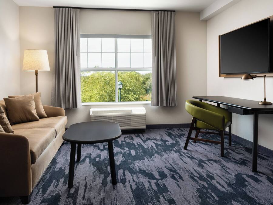 A modern hotel room with a sofa, a small table, a desk with a chair, a flat-screen TV, and a large window with curtains.