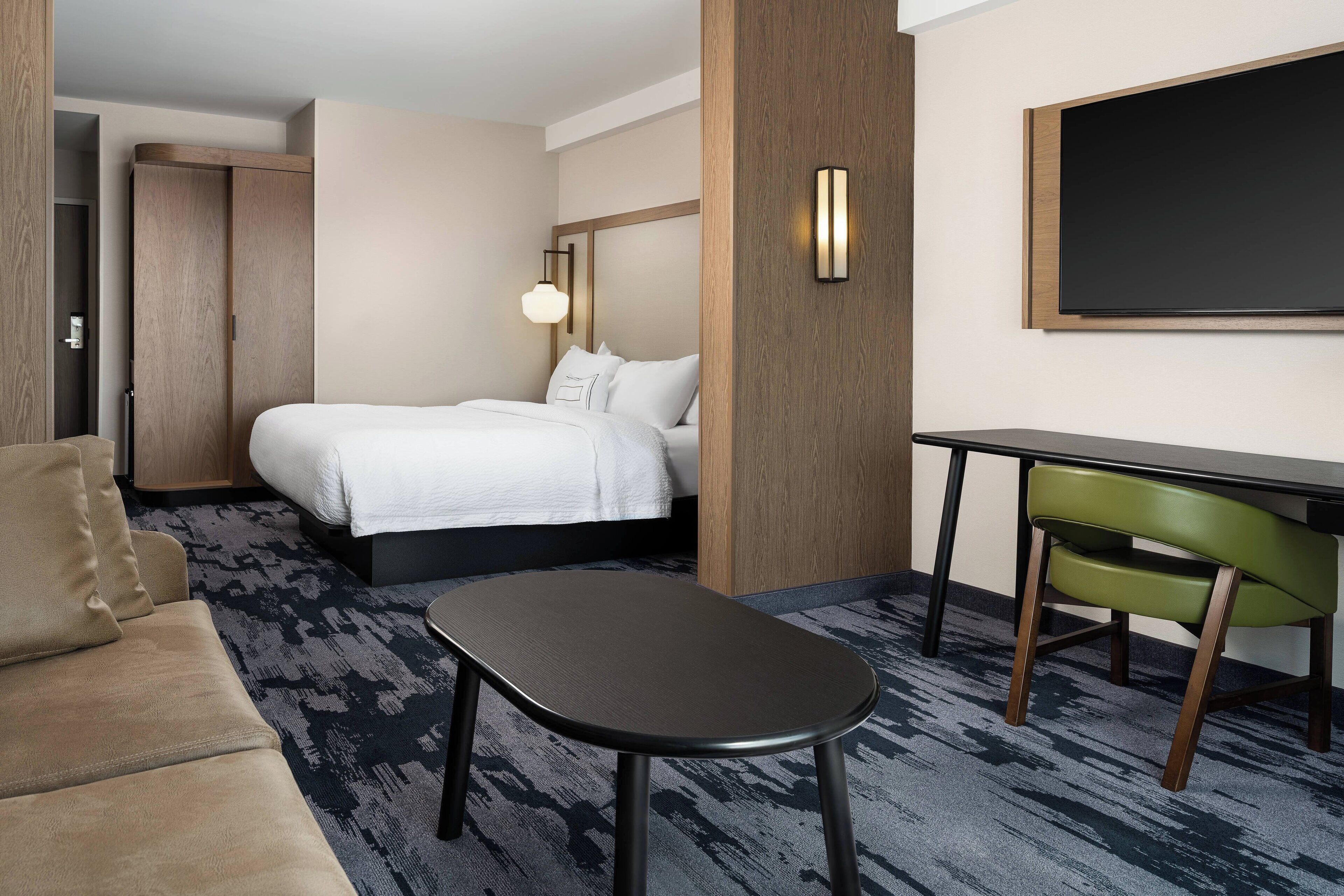 Una habitación de hotel con cama, escritorio, sillón verde, sofá y televisión montada en la pared.