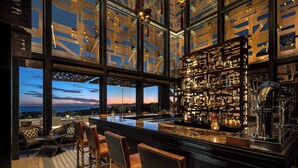 A modern bar with a view of the sunset, featuring a bar counter, seating area, and a well-stocked shelf with bottles. - Zadun Los Cabos, a Ritz-Carlton Reserve (San José del Cabo)