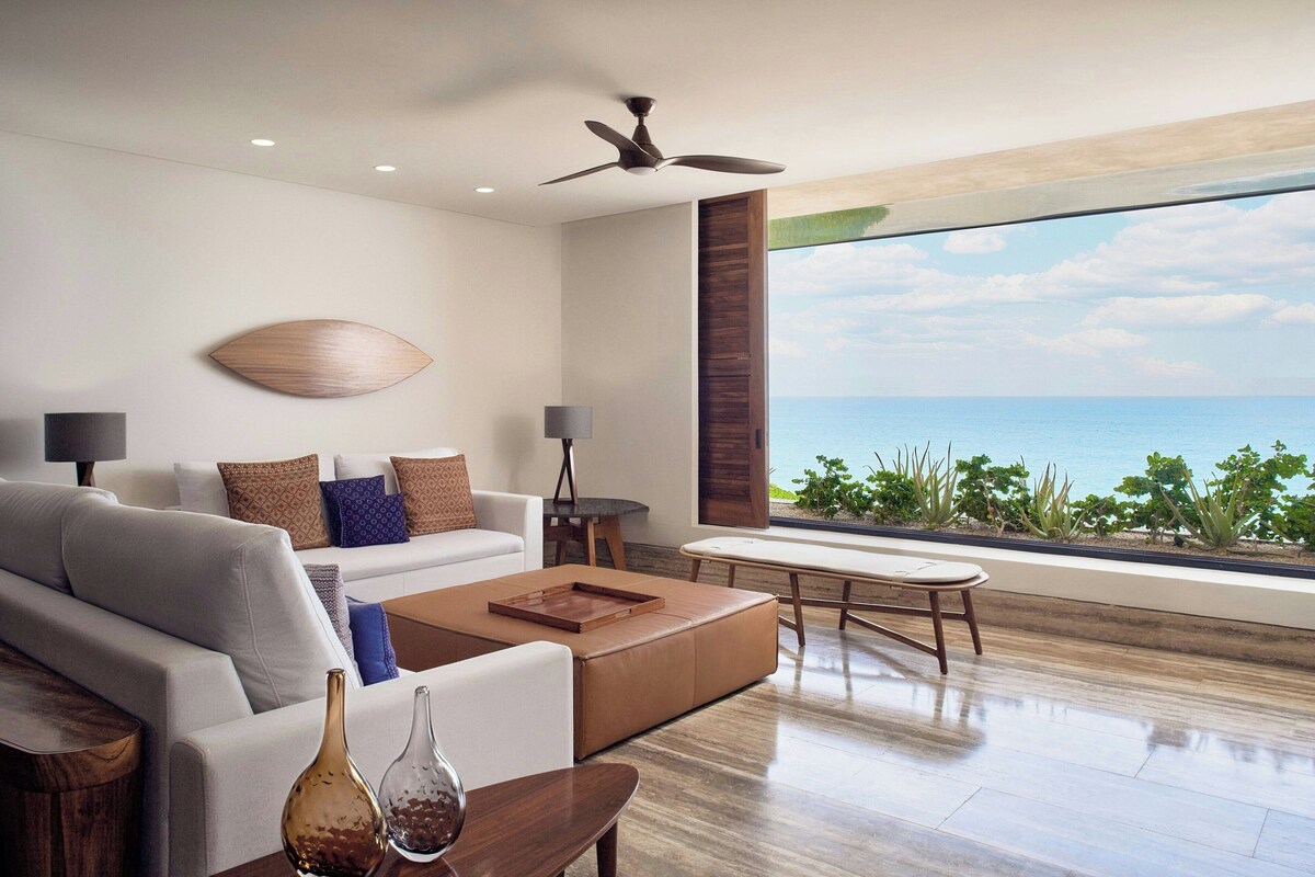A modern living room with a large window overlooking the ocean, a ceiling fan, and a minimalist design.