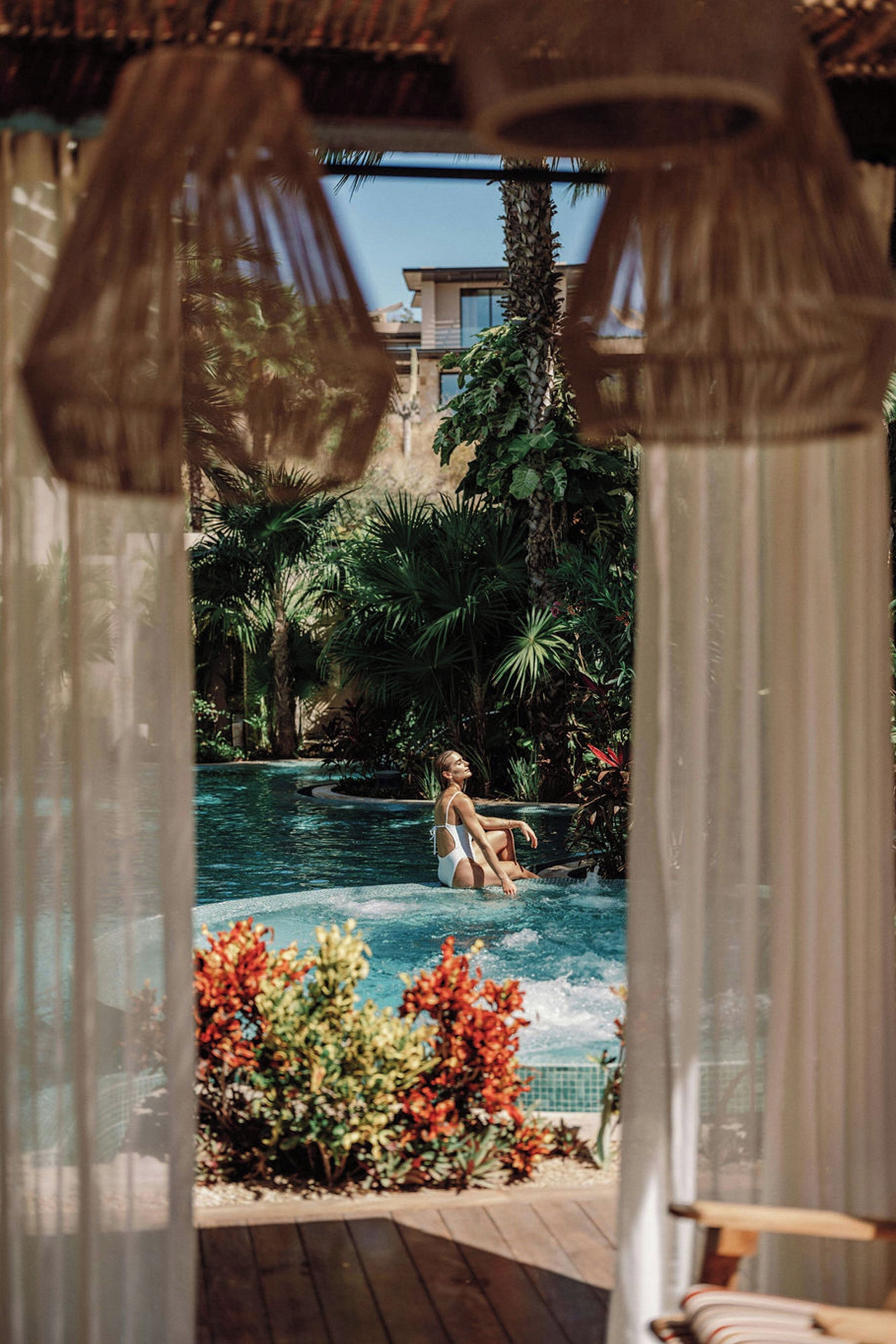 Zona de piscina amb una persona asseguda en una cadira relaxant, envoltada de plantes tropicals i una piscina blava cristal·lina.