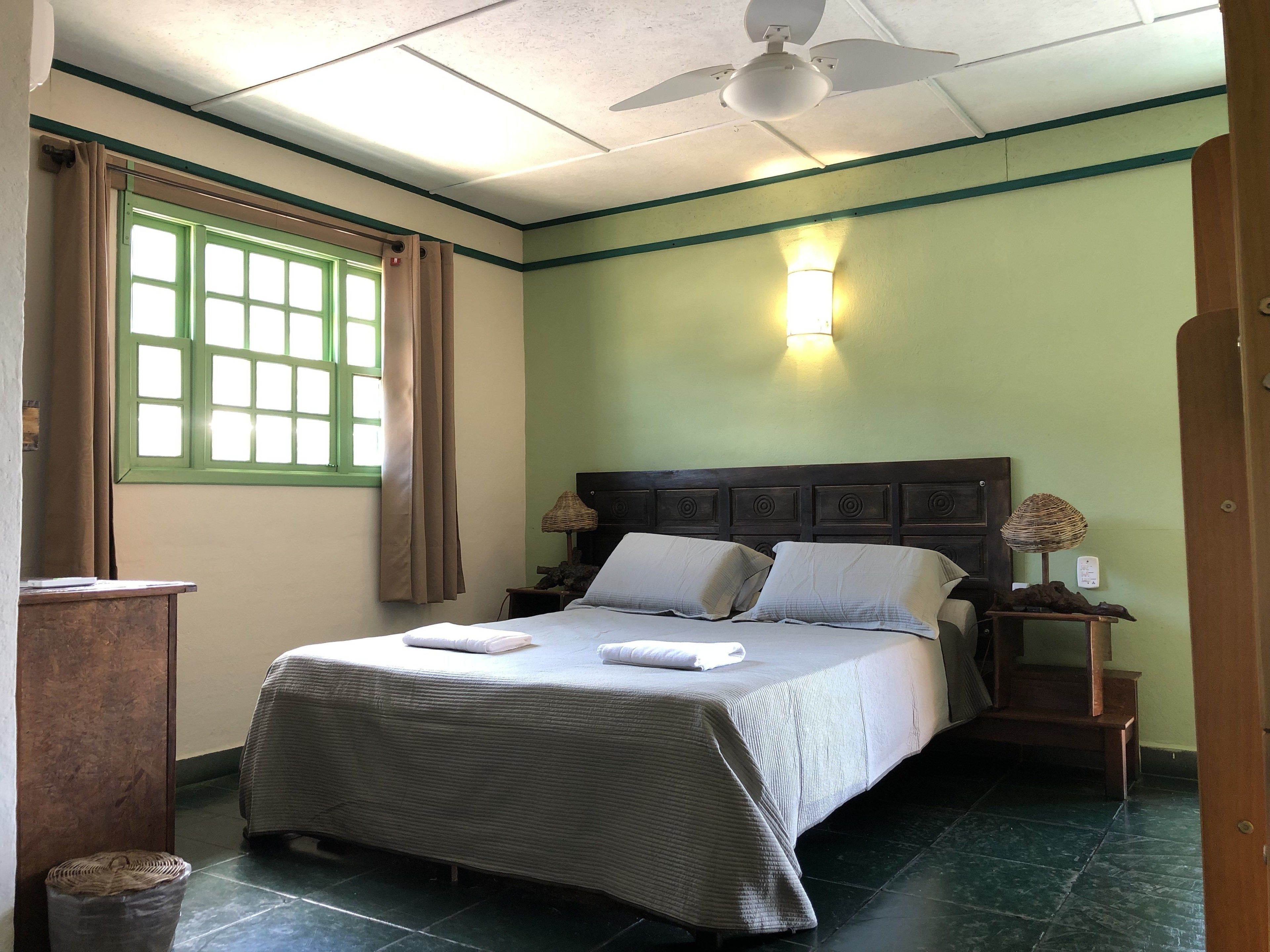 Una cama bien tendida con almohadas y una toalla doblada, una lámpara de noche, una ventana con cortinas y un ventilador de techo.