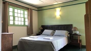Una cama bien tendida con almohadas y una toalla doblada, una lámpara de noche, una ventana con cortinas y un ventilador de techo.