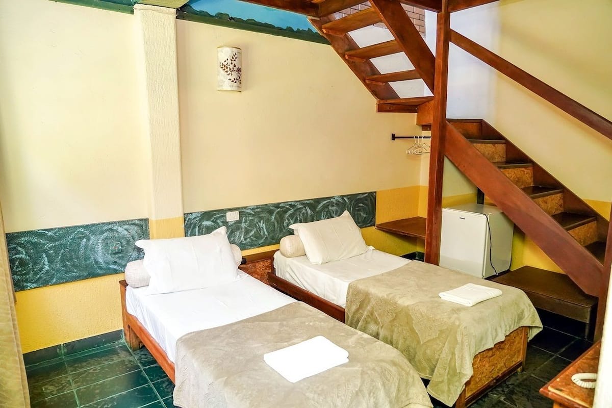 A hotel room with two single beds, a small desk, a chair, a staircase, and a wall-mounted light fixture.