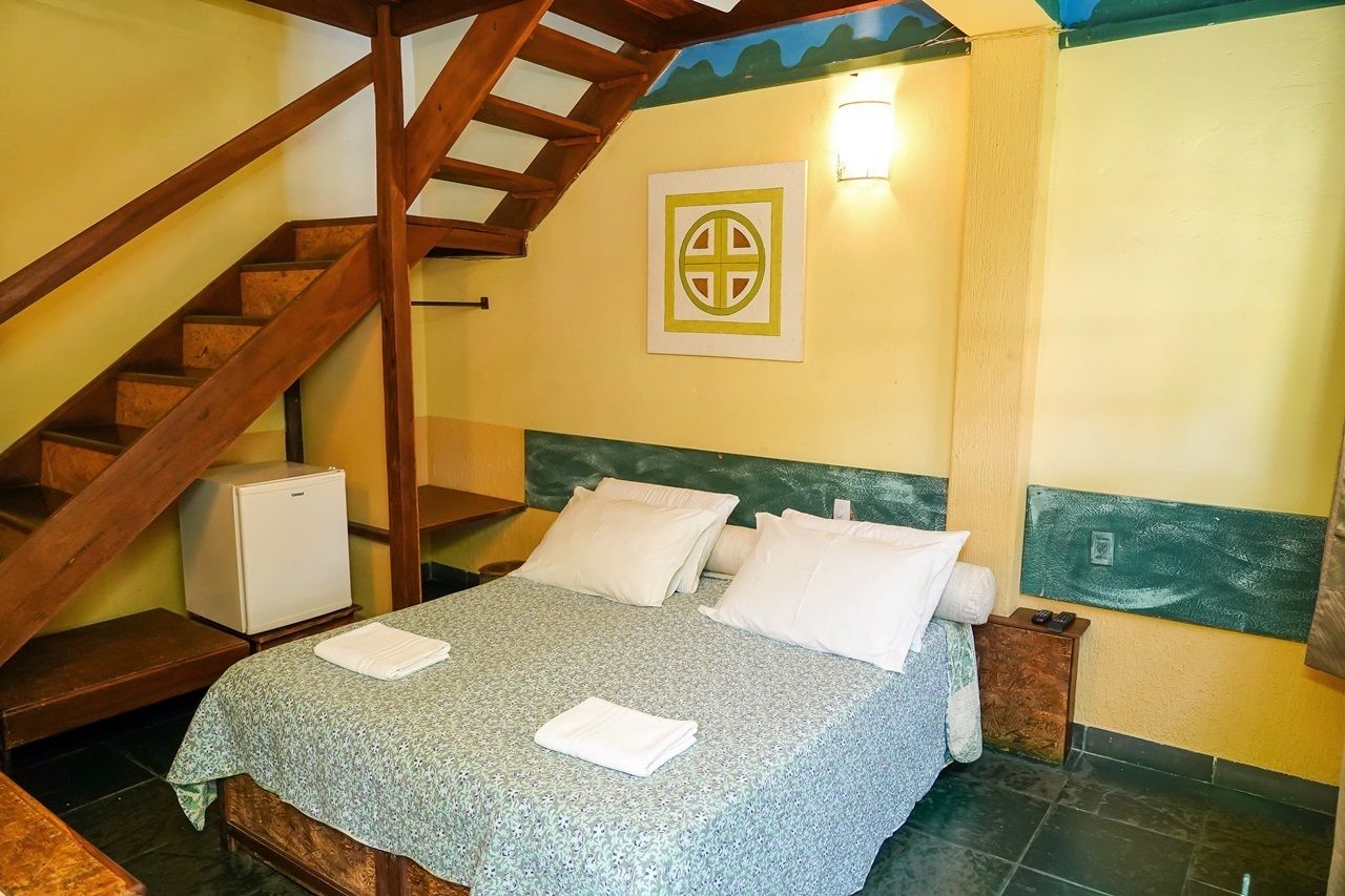 A bedroom with a bed, pillows, a small bedside table, a nightstand, a wall-mounted light, and a staircase.
