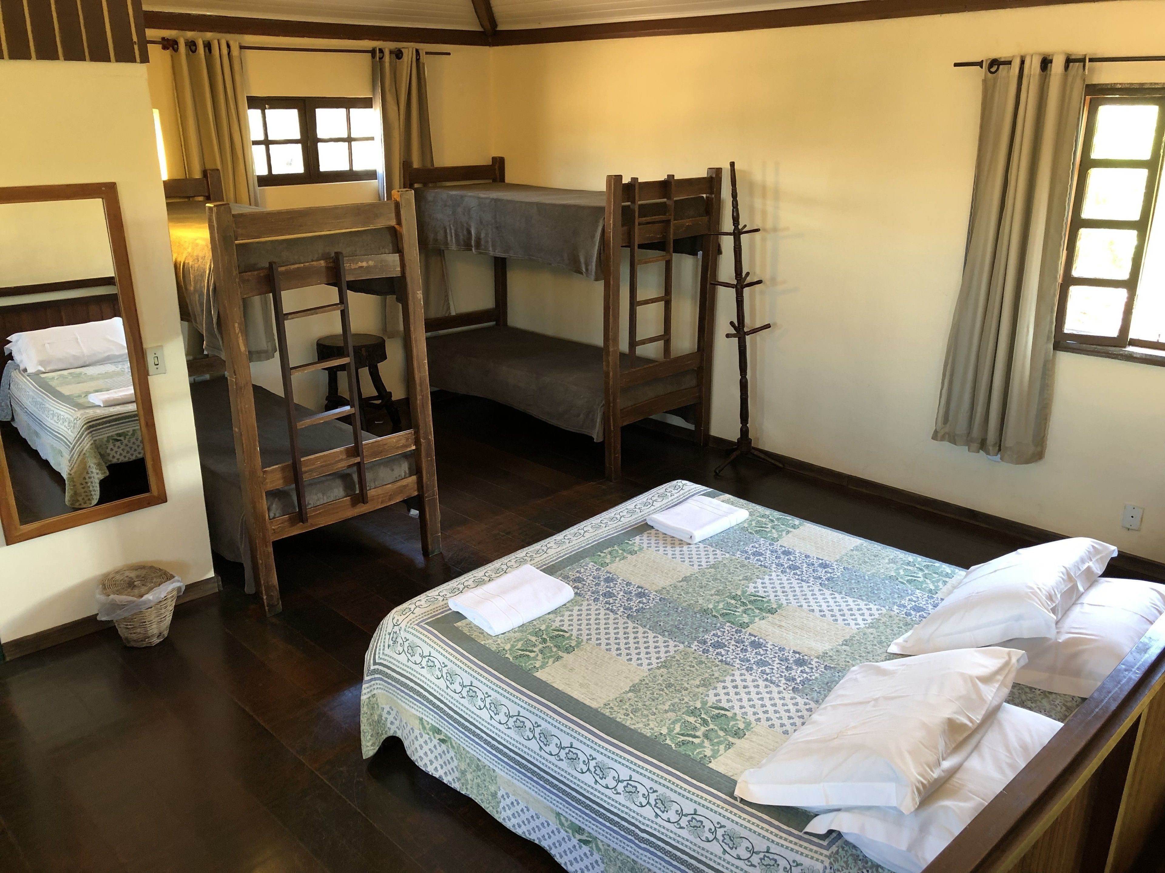 Ein Zimmer mit Etagenbett, Einzelbett, einer Holzleiter, einem kleinen Tisch, einem Spiegel und einem Fenster mit Vorhängen.