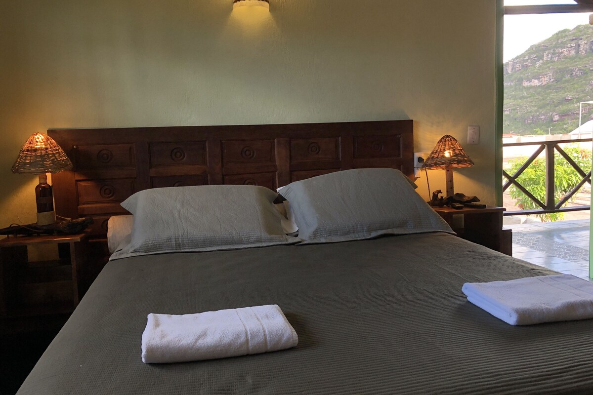 A neatly made bed with pillows and folded towels, two bedside lamps, and a view of a mountainous landscape through a balcony.