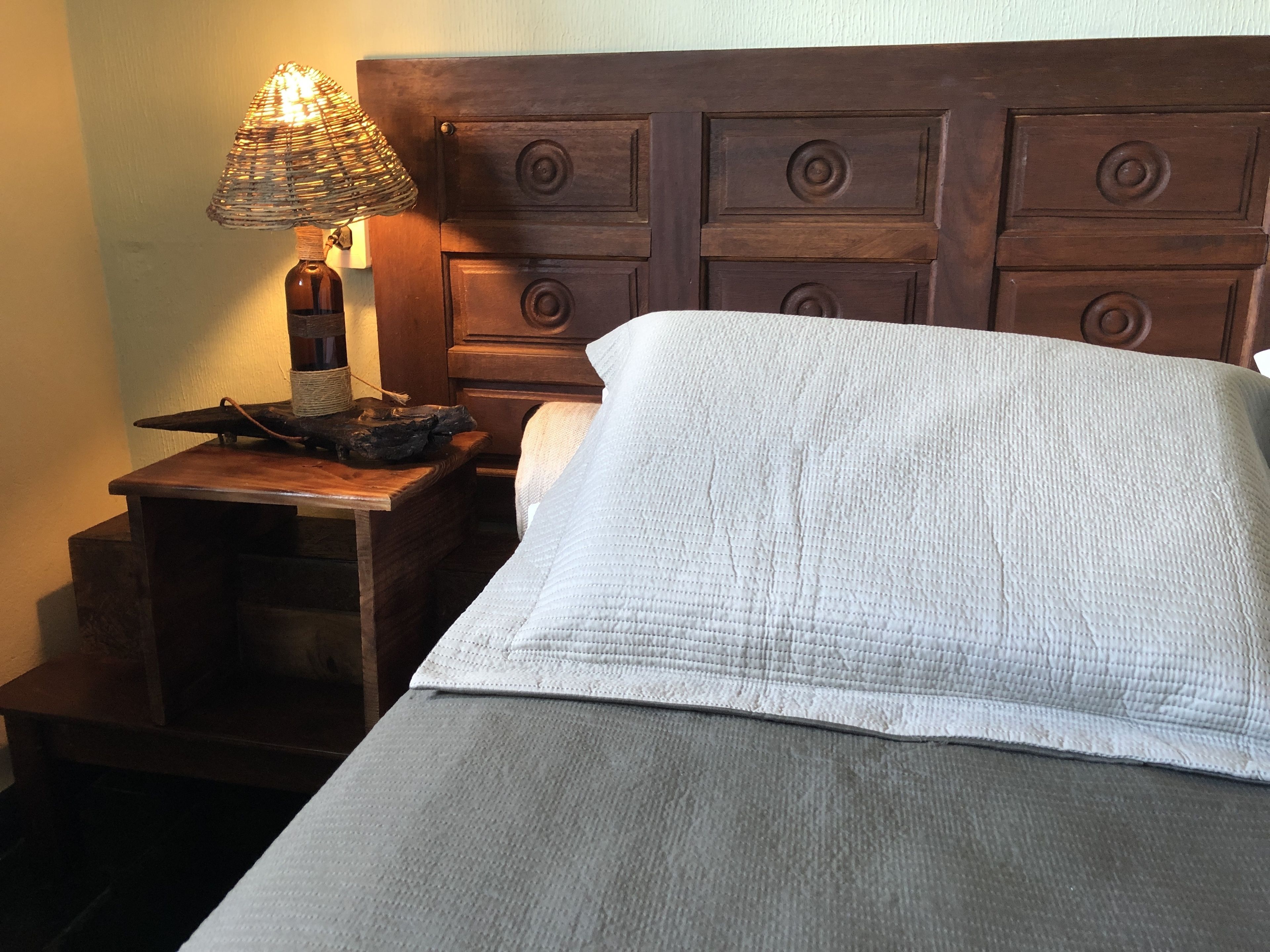 A bed with a white quilt, a wooden nightstand with a lamp, and a wooden headboard.