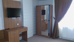 Ein Zimmer mit Schreibtisch aus Holz, einem an der Wand befestigten Fernseher, einem Kleiderschrank mit Spiegel und einem Fenster mit Vorhängen.